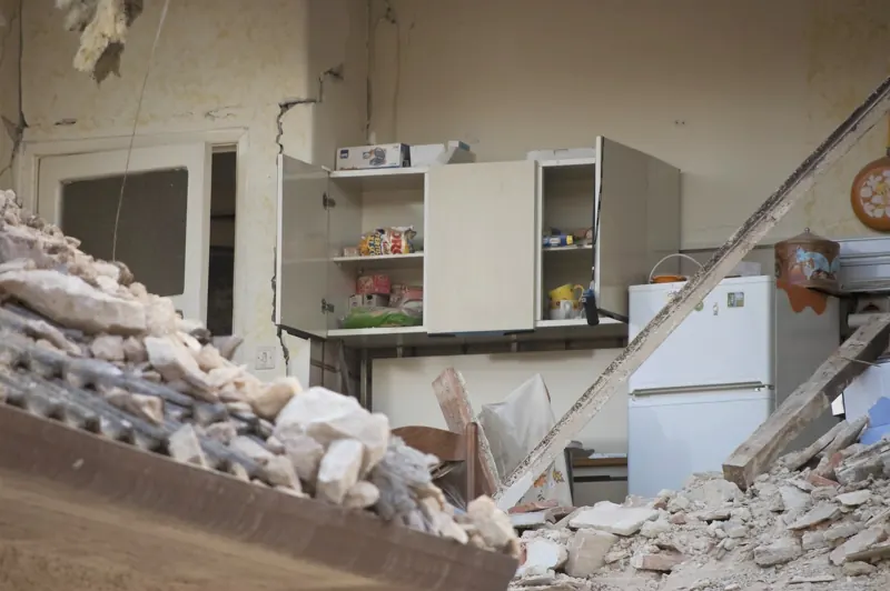 A kitchen with a collapsed wall and a cupboard hanging open after an earthquake